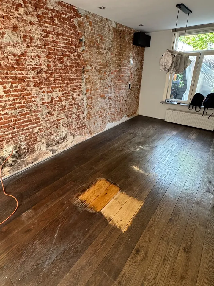 Donkere, versleten eiken plankenvloer naast een kale bakstenen muur, met midden in de kamer één klein vierkant teruggeschuurd tot licht hout