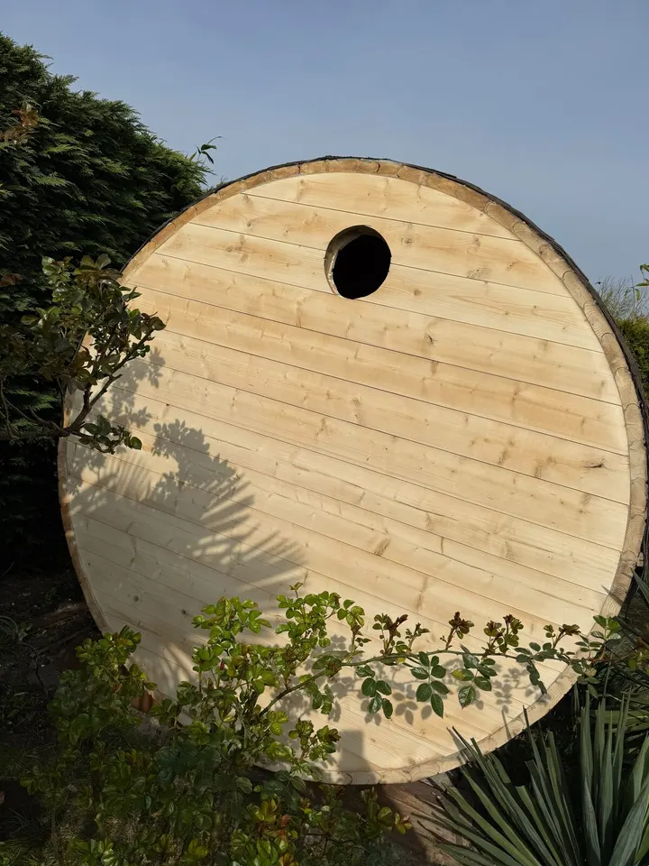 Het ronde eind van een tonvormig tuinhuisje in kale, lichte planken, met een klein rond raampje