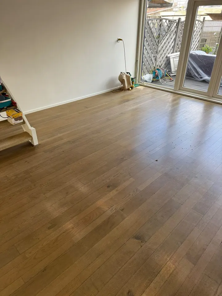 Eiken strokenvloer in een woonkamer met tuindeuren, dof en met plekken, met gereedschap tegen de muur