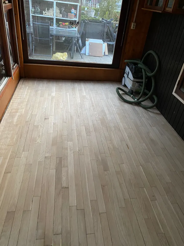 Strokenvloer in de tuinkamer teruggeschuurd tot kaal, egaal licht hout, met de stofafzuiging nog in de hoek