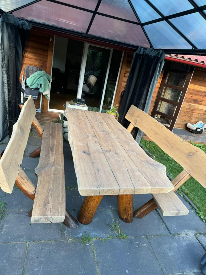 Een rustieke tuintafel en twee banken teruggeschuurd tot ruw hout onder een tuinoverkapping