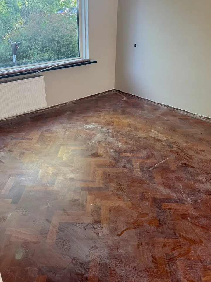 Visgraatparket onder een vlekkerige, witachtige laag oude afwerking, in een kamer met een groot raam naar de tuin