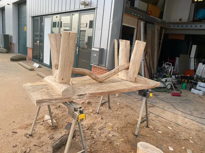 Het boomstamonderstel van een tafel ondersteboven op schragen buiten de werkplaats, twee paar poten verbonden door een gebogen tak