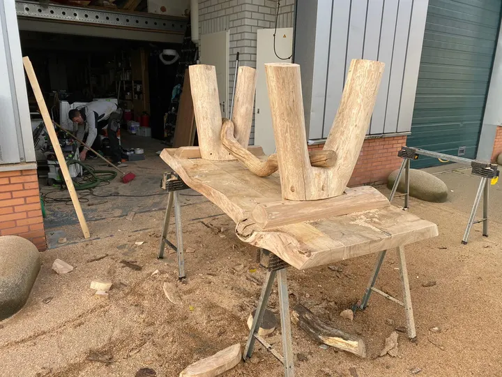 Hetzelfde boomstamonderstel op schragen buiten de werkplaats, met een vakman aan het werk in de deuropening erachter