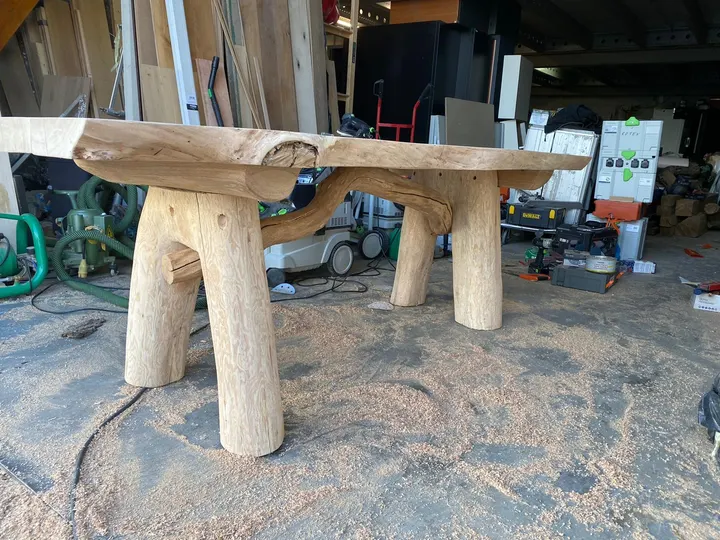Een boomstamtafel met natuurlijke rand op boomstampoten, geschuurd tot ruw licht hout, in de werkplaats tussen het zaagsel
