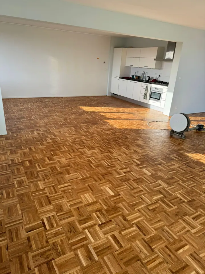 Grote open vloer van eiken mozaïekparket die van de woonkamer doorloopt naar een witte keuken, met een kantenschuurmachine aan de muur