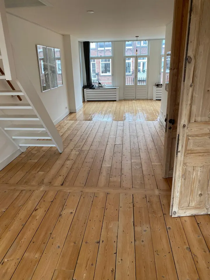 Brede grenen vloerdelen die door een woonkamer in een grachtenpand lopen, langs een open witte trap naar hoge voorramen