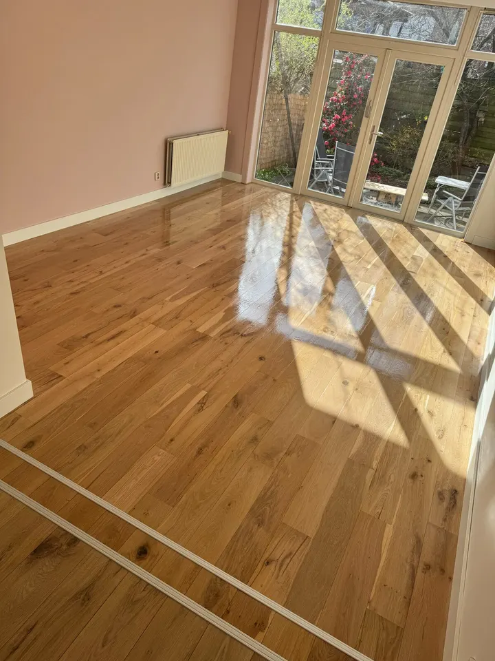Dezelfde eiken vloer net behandeld, nog nat en glanzend, met het zonlicht van de tuindeuren erin weerspiegeld