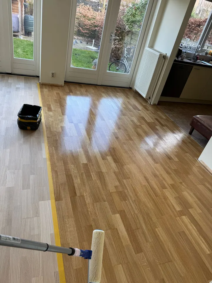 Een eiken strokenvloer verdeeld door een lijn afplaktape, ruw geschuurd hout aan de ene kant en vers gelakt hout aan de andere, roller op de voorgrond