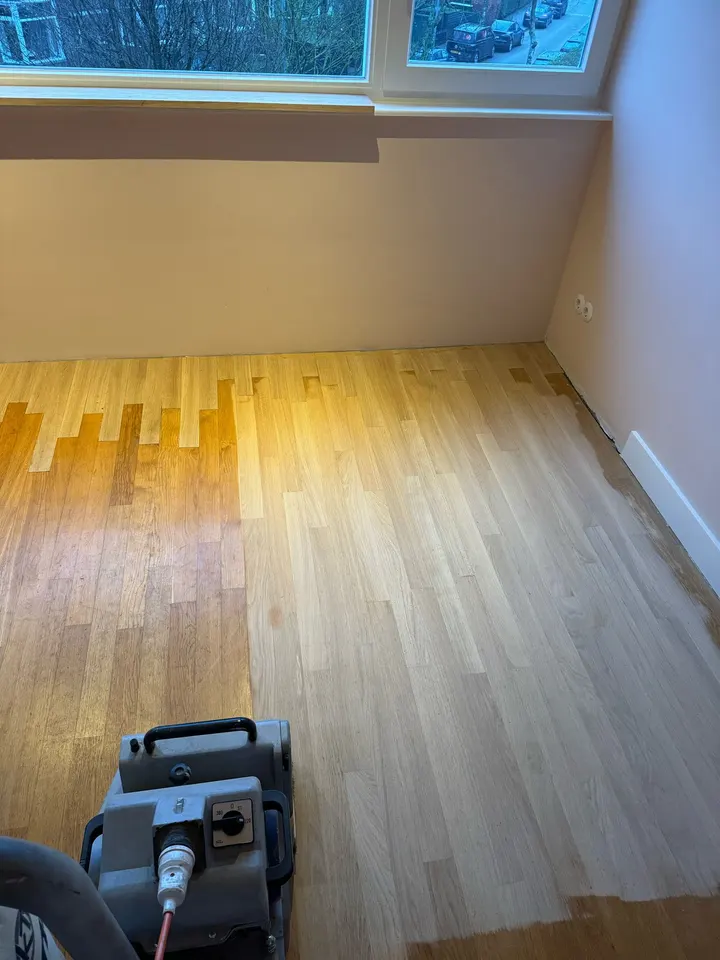 Een eiken strokenvloer half geschuurd, licht nieuw hout aan de ene kant van de lijn en vergeelde oude lak aan de andere, bandschuurmachine op de voorgrond