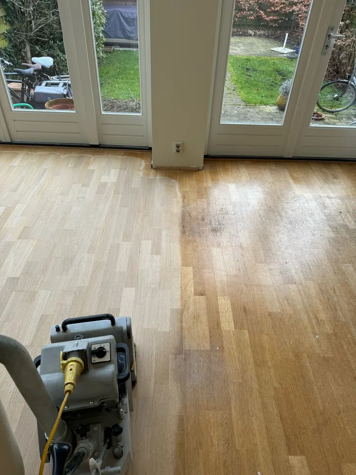Een eiken strokenvloer die bij de tuindeuren wordt geschuurd, licht geschuurd hout links en donkerder, verkleurd hout dat rechts nog moet komen